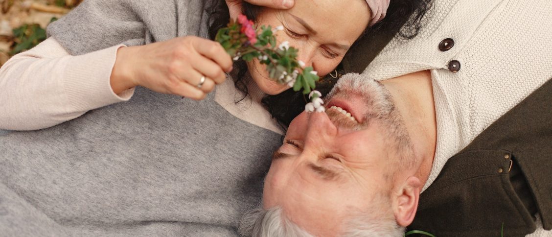 From above of cheerful senior wife wearing wide scarf and headband with flower bouquet in hand and happy elderly gray haired husband in warm clothes lying on ground with fallen leaves in park with closed eyes