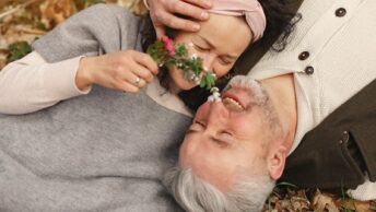 From above of cheerful senior wife wearing wide scarf and headband with flower bouquet in hand and happy elderly gray haired husband in warm clothes lying on ground with fallen leaves in park with closed eyes