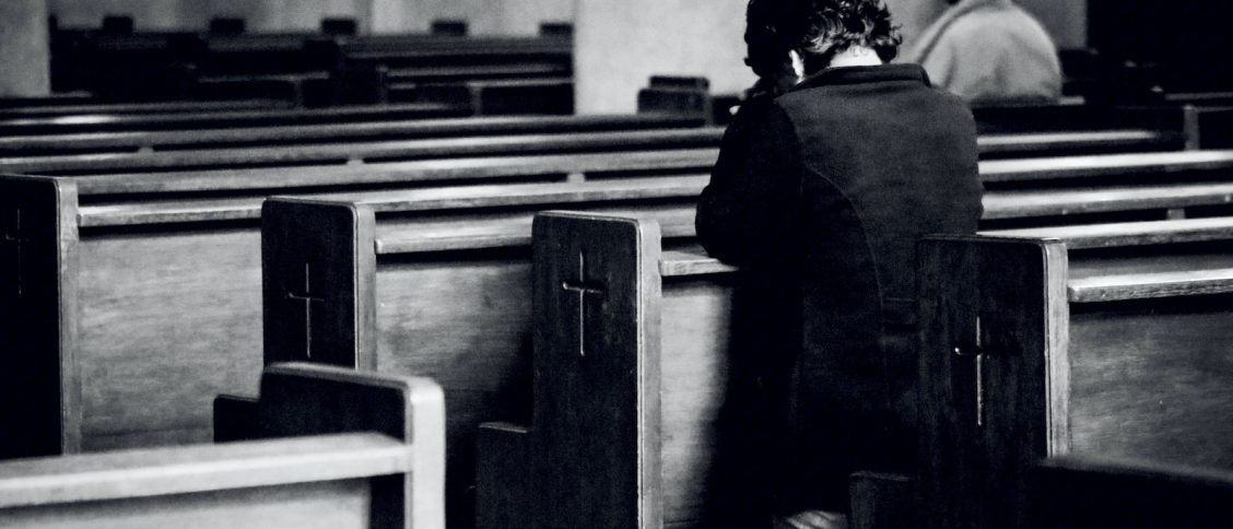 Black and white back view of anonymous male believers standing on knees near wooden benches with symbol of cross on surface during mass in cathedral