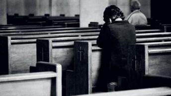 Black and white back view of anonymous male believers standing on knees near wooden benches with symbol of cross on surface during mass in cathedral