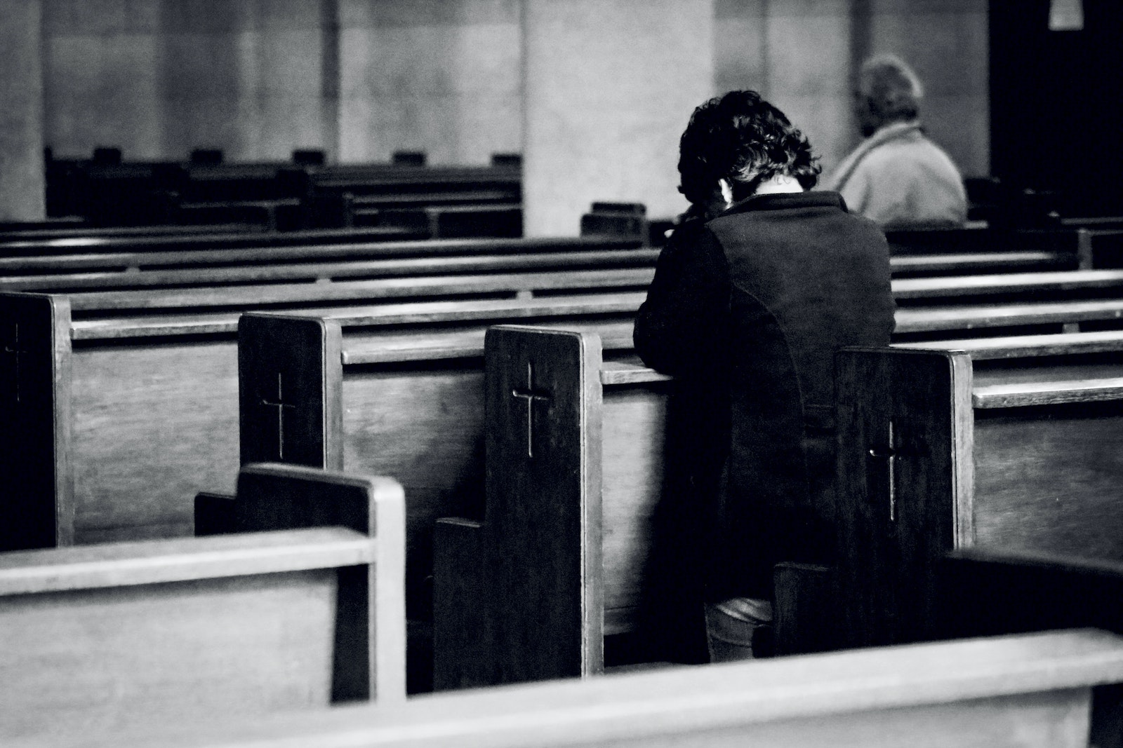 Black and white back view of anonymous male believers standing on knees near wooden benches with symbol of cross on surface during mass in cathedral
