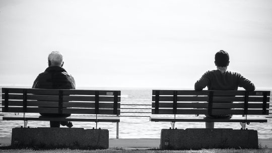 Grayscale Photo 2 Person Sitting in a Separate Benches on the Seaside