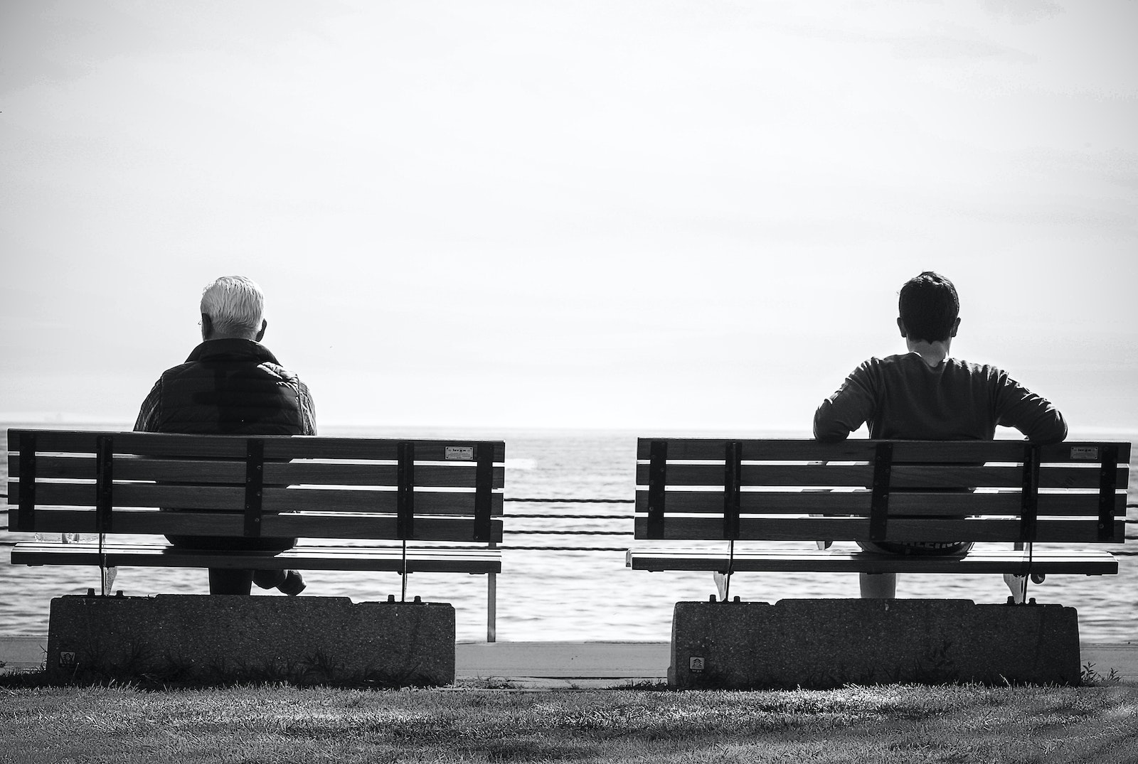 Grayscale Photo 2 Person Sitting in a Separate Benches on the Seaside