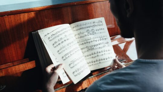 Back view of crop anonymous male pianist watching music paper with written notes near piano